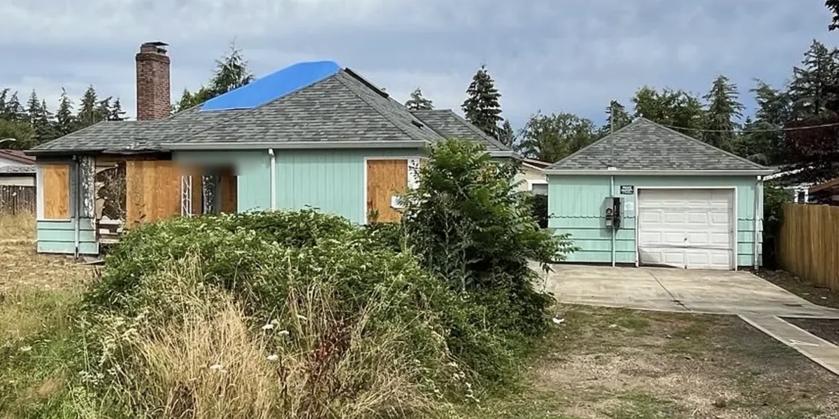 The same home before, with a blue tarp over a worn gray roof, boarded-up windows, and mint-green siding