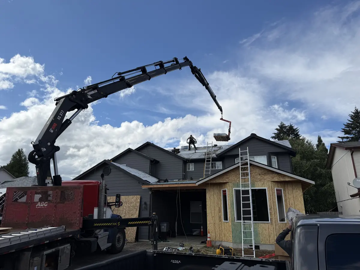 Boom truck lifting a pallet of shingles onto the roof of a two-story gray home in Vancouver, WA while a Big Bear Exteriors roofer works on the roof
