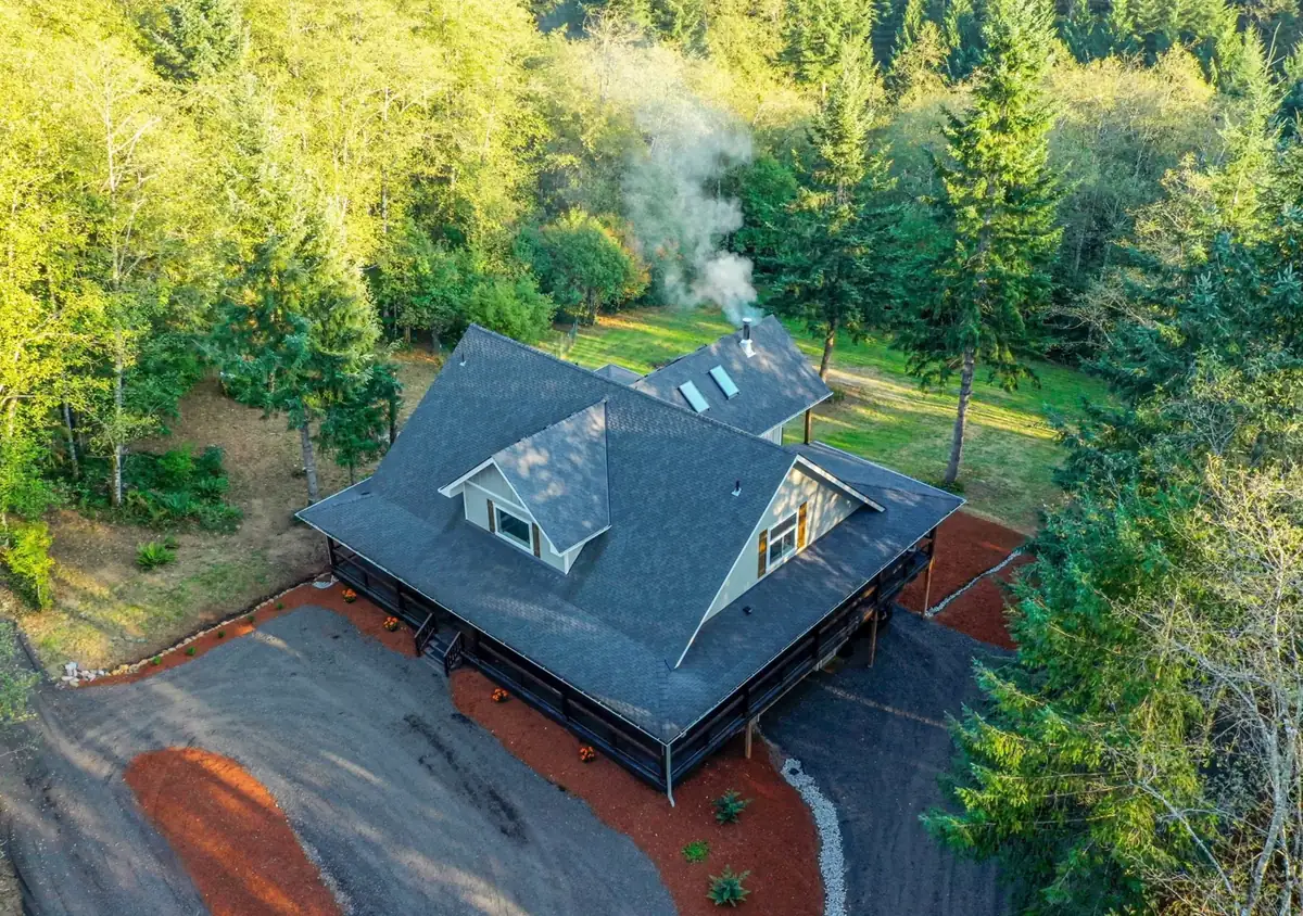 Aerial view of a rural Battle Ground, WA home with a new charcoal-gray shingle roof, two gable dormers, skylights, and a wraparound porch, surrounded by tall firs